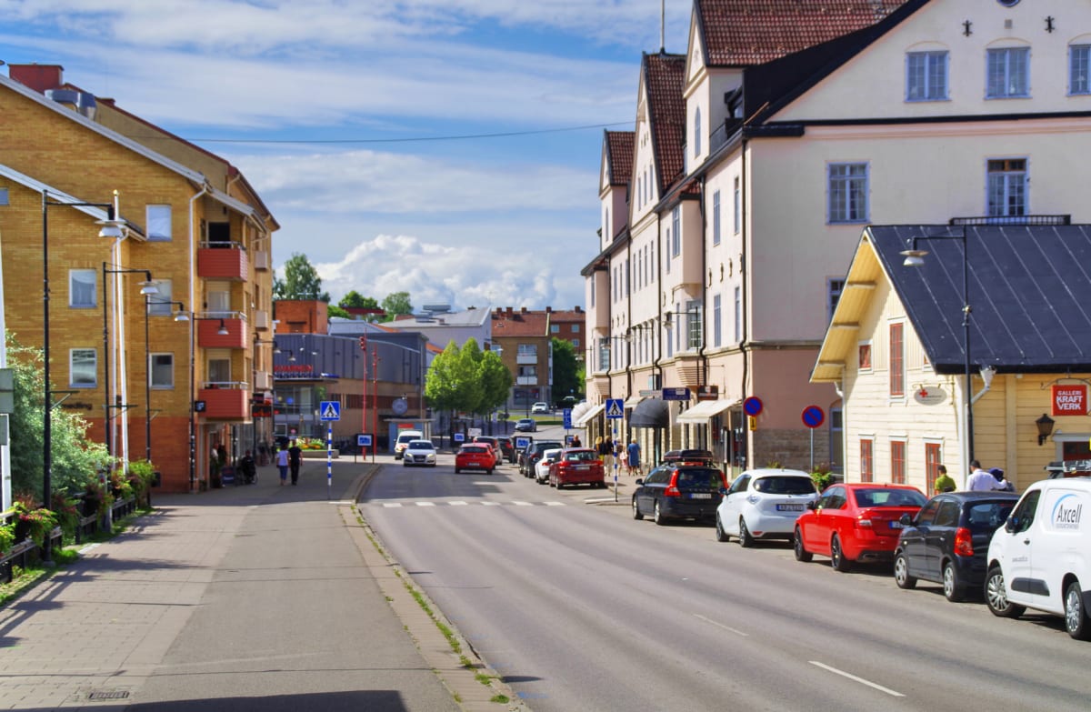 En gata i en stad på en solig dag, kantad av byggnader i olika färger som gult och rosa. Flera bilar är parkerade längs gatan, och en del människor går på trottoarerna. Blå himmel med vita moln syns ovanför byggnaderna.