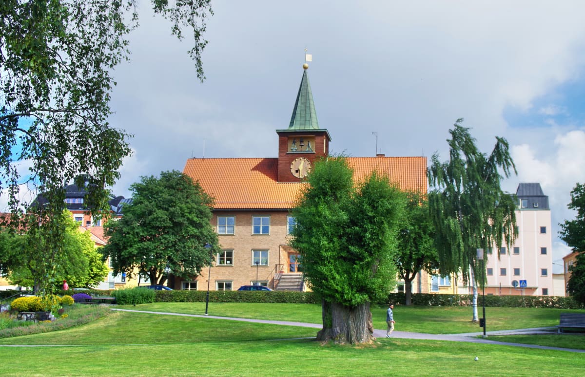 En bild som visar en gräsplätt i förgrunden, med träd och en byggnad bakom. Byggnaden har ett tegeltak och ett grönfärgat torn med klocka. På gräsplätten syns en person på en gångväg intill ett träd.