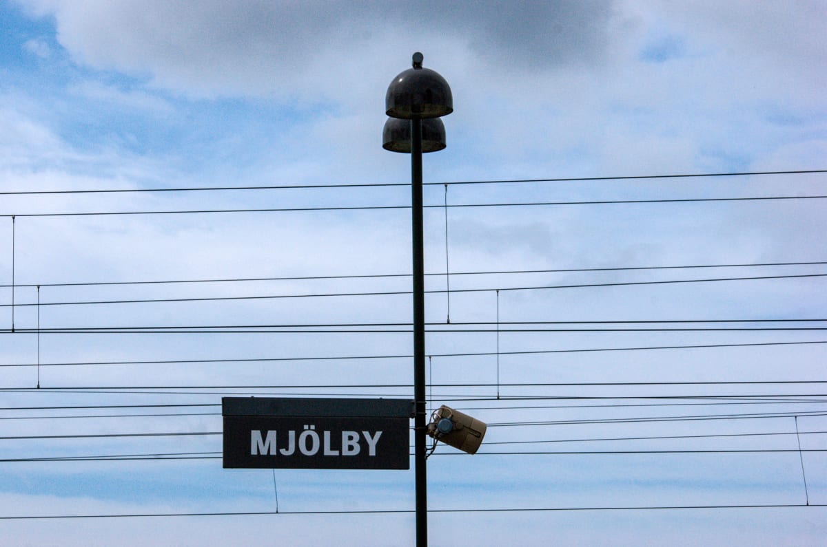 En bild tagen underifrån visar en stolpe med två klockformade objekt överst och en skylt med texten "MJÖLBY" fäst på den. Flera horisontella ledningar spänns över bilden, med en kamera eller belysningsarmatur fäst vid stolpen.