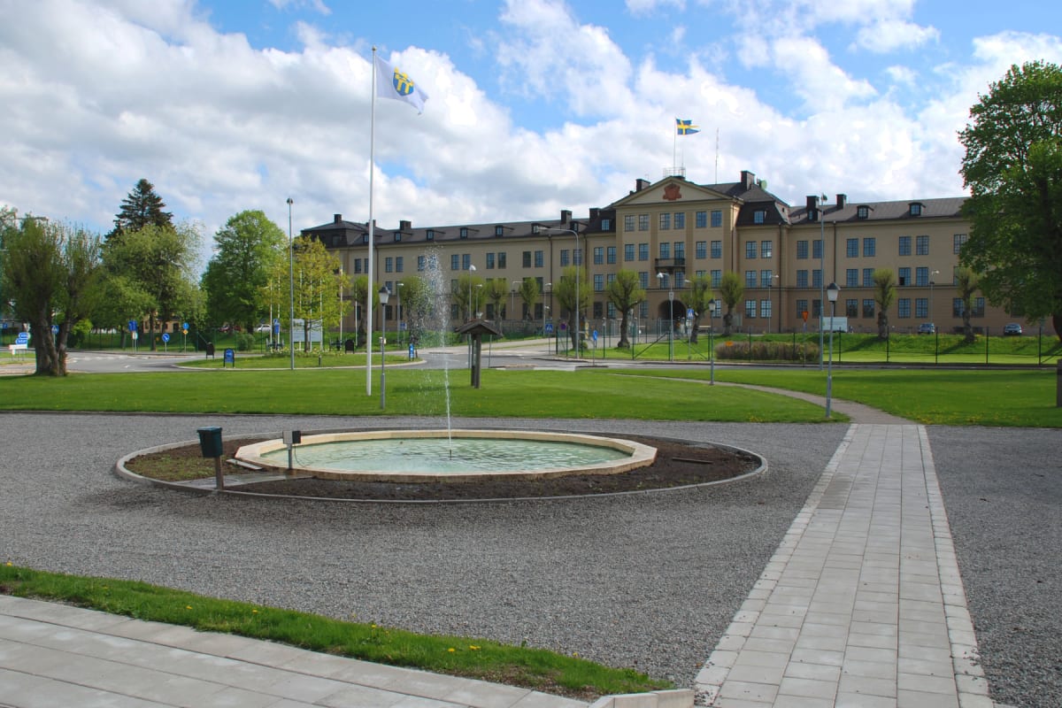 En bild visar ett torg med en fontän i förgrunden. I bakgrunden syns en stor, ljus byggnad med en svensk flagga på taket och en flagga med ett vapen. Runt fontänen finns gräsytor, grusgångar och träd, och en blåsig molnig himmel.