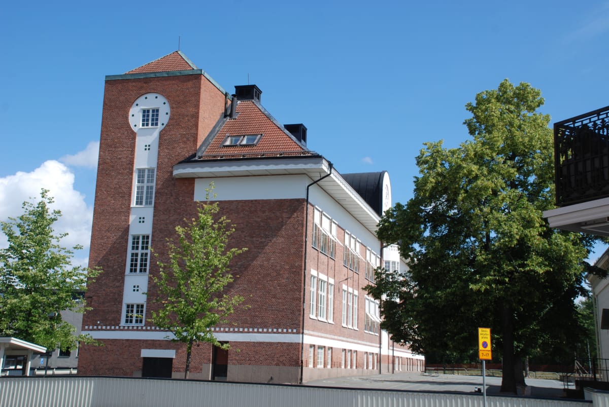En stor, röd tegelbyggnad med vita detaljer under en klarblå himmel. Byggnaden har ett torn med ett runt fönster och många kvadratiska fönster med vita karmar. Framför byggnaden syns flera gröna träd och en ljusbeige mur. Längst till höger syns en gu