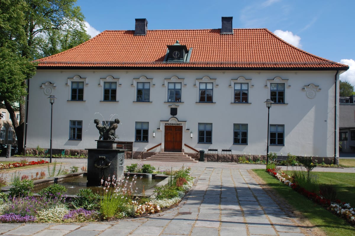 En vit byggnad med rött tegeltak och en klocka högst upp, framför vilken det finns en fontän med statyer av keruber. En stengång leder fram till byggnadens entré, omgiven av blomsterplanteringar och gräsmattor under en klarblå himmel.