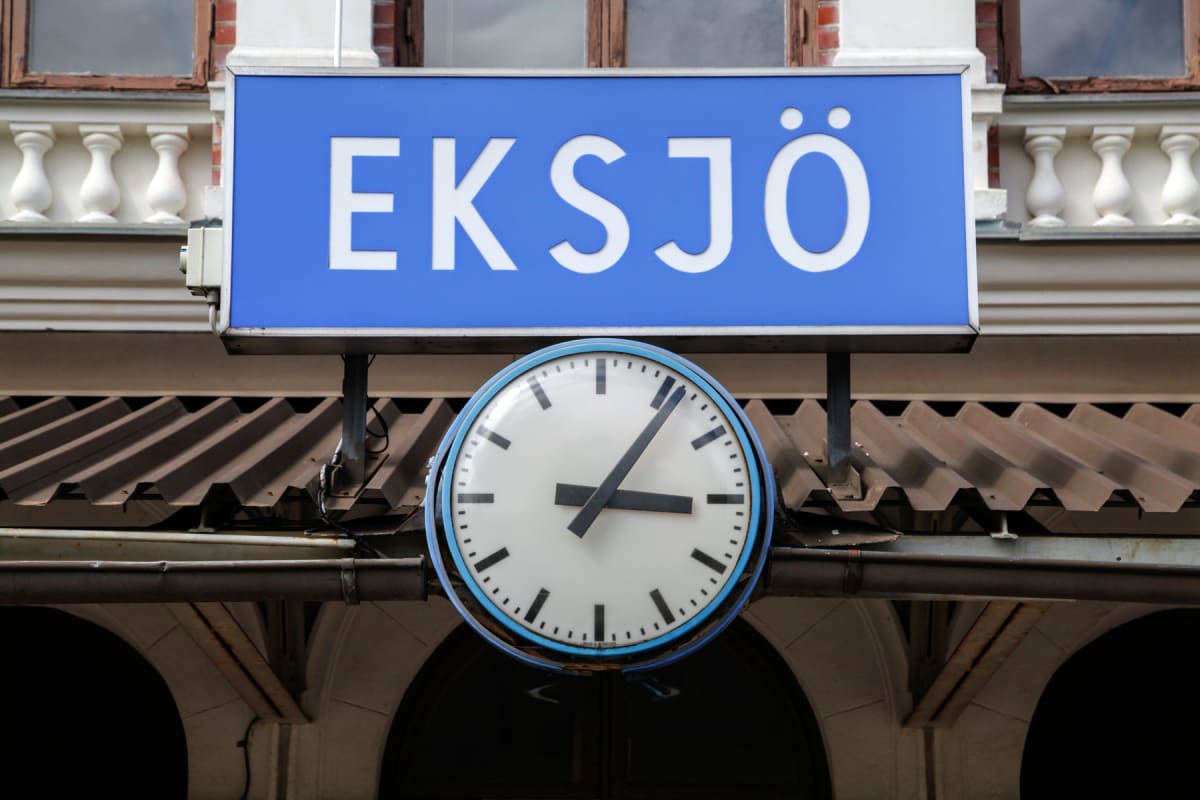 En blå skylt med texten "EKSJÖ" i vitt hänger ovanför en rund, vit stationsklocka med svarta visare och markeringar. Bakom skylten och klockan syns en del av en byggnad med fönster och en takkonstruktion, samt en mörkblå himmel.