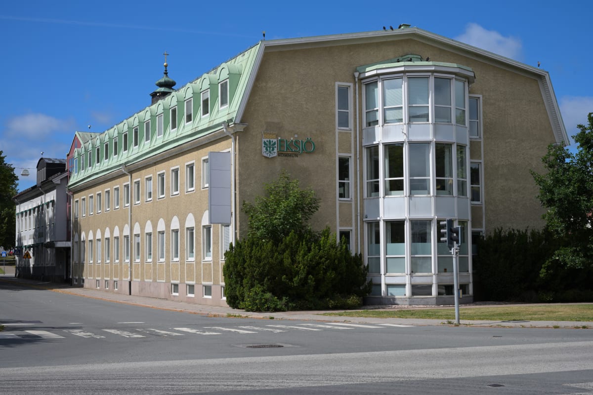 En gulbeige byggnad med grönt plåttak och många fönster. Byggnaden har en utskjutande glasfasad. Framför byggnaden finns en gata med ett övergångsställe och buskar. Himlen är klarblå med några vita moln. En trafiklampa syns vid sidan av byggnaden.