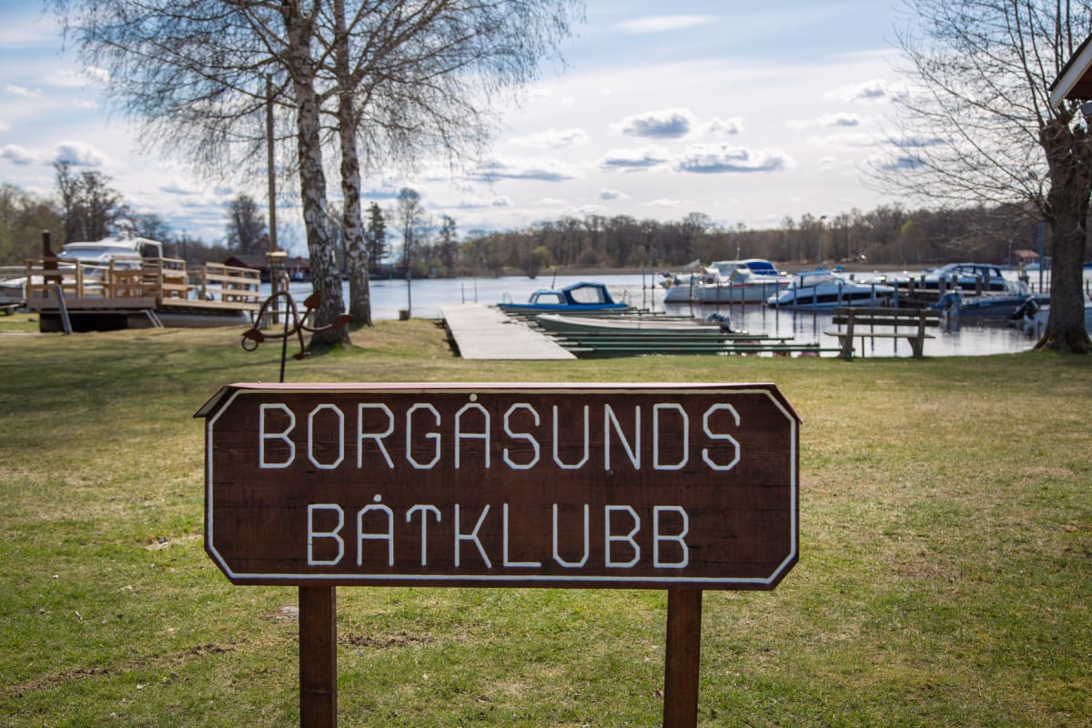 En brun skylt med texten "BORGÅSUNDS BÅTKLUBB" i vitt står i förgrunden på en grön gräsmatta. I bakgrunden syns en brygga med flera förtöjda båtar vid ett vattendrag, omgivet av träd och himmel med vita moln.