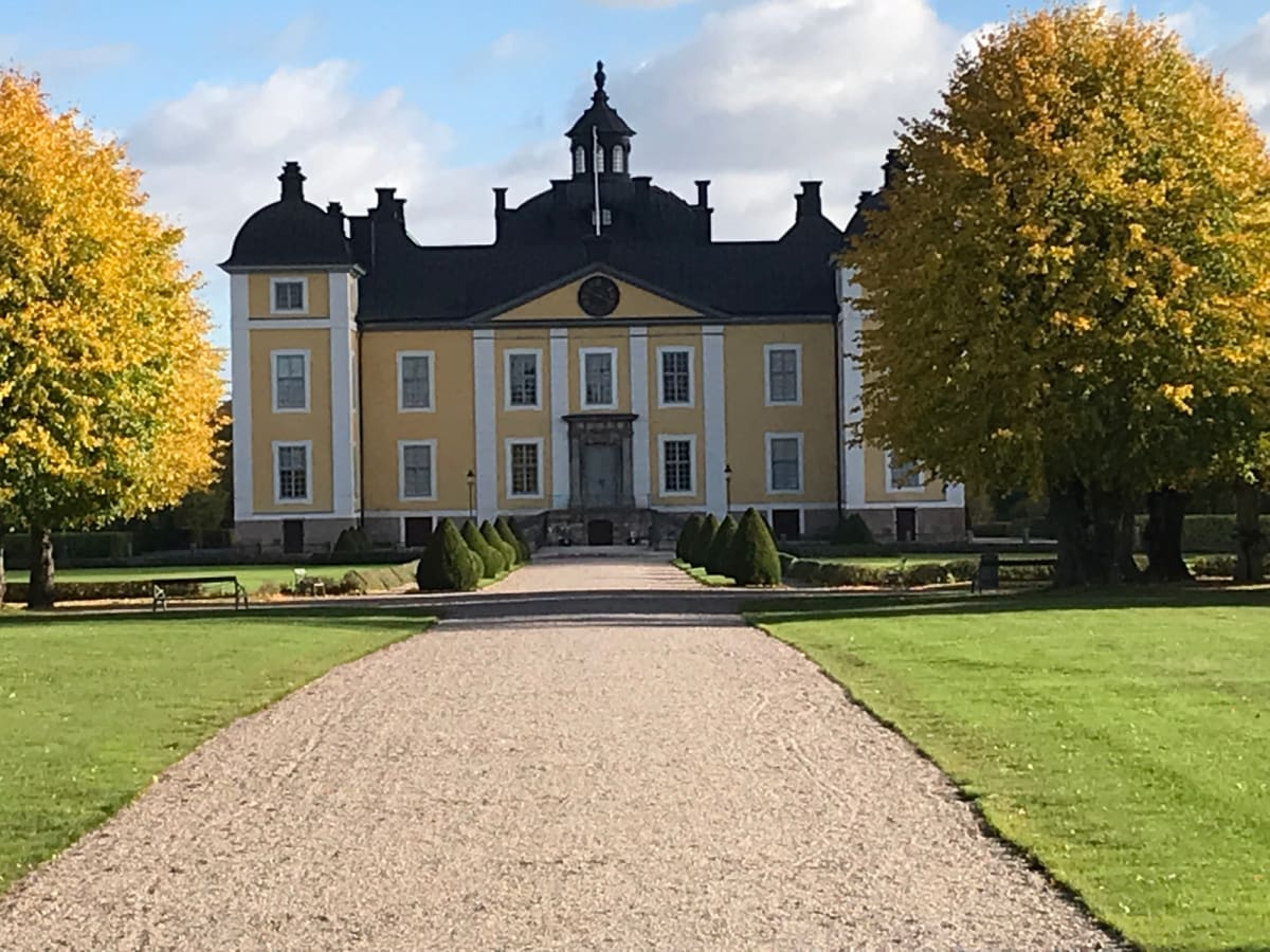 En stor, gul byggnad med många fönster och ett mörkt tak utgör bildens centrum. En grusgång leder fram till byggnaden, flankerad av gräsmattor och mindre buskar. På vardera sida om byggnaden står träd med gulgröna löv under en blå himmel med vita mol