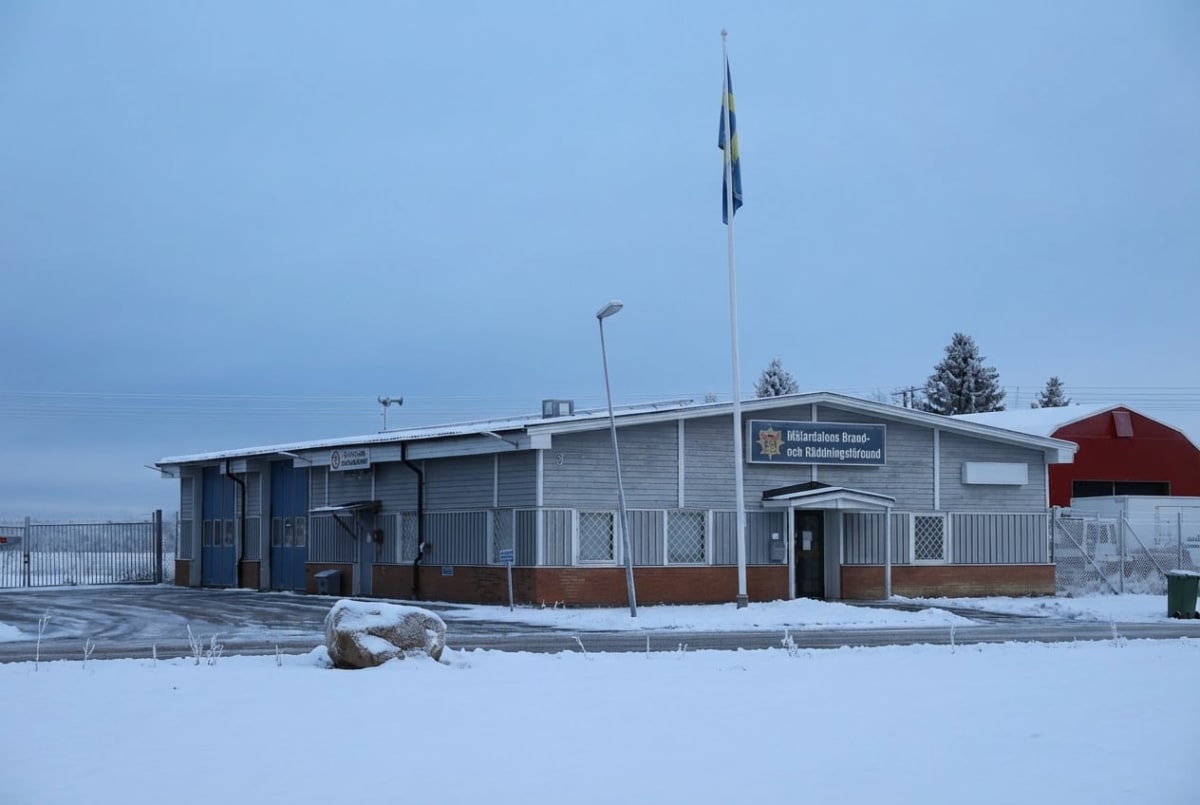 En stor, ljusgrå byggnad med skylten "Mälardalens Brand- och Räddningsförbund" står i fokus på en snötäckt mark. En flaggstång med en svensk flagga är placerad framför byggnaden. I bakgrunden syns ett rött tak, snötäckta träd och en grå himmel med öv
