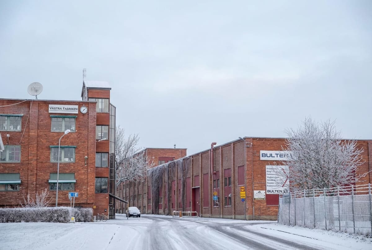 En vinterbild av en industrimiljö med snötäckta vägar och byggnader. De tegelröda byggnaderna har vita skyltar, bland annat "VÄSTRA FABRIKEN" och "BULTEN". Ett fordon står parkerat på vänster sida av vägen och träd samt buskar är täckta av rimfrost.