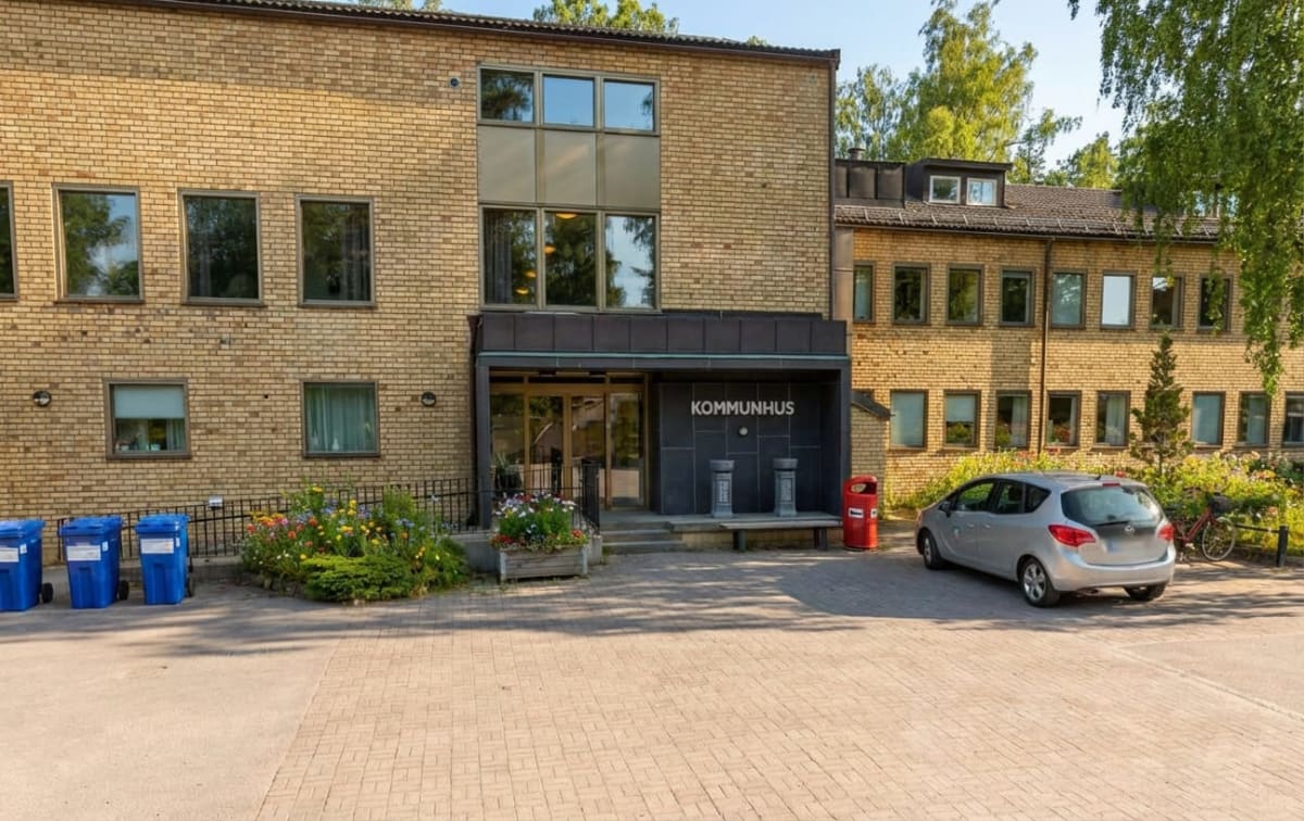 En ljusgul tegelbyggnad med många fönster och en entré som skyltas "KOMMUNHUS" framifrån. Framför entrén står en silverfärgad bil parkerad till höger, och till vänster står tre blåa sopkärl. Blommor och grönska syns vid byggnadens bas.