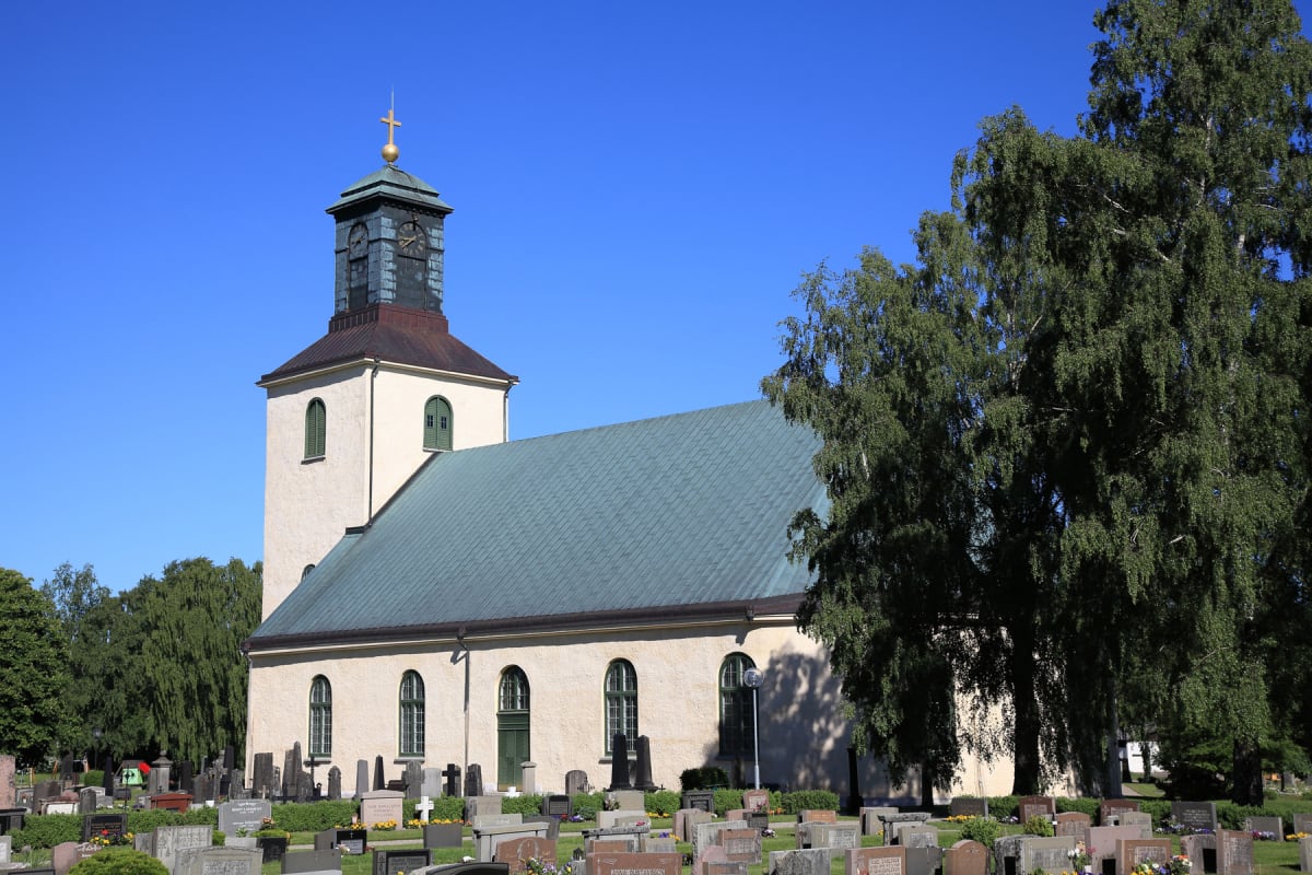 En beige kyrka med grönt tak och ett klocktorn med guldryck upptill står omgiven av en kyrkogård med gravstenar och grönskande träd under en klarblå himmel.