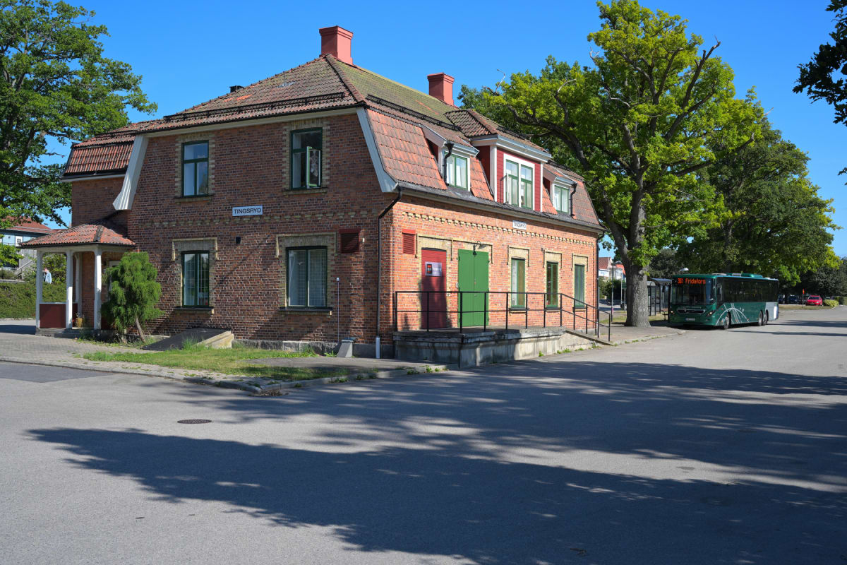 En röd tegelbyggnad med tegeltak och röds- och grönmålade dörrar. En grön buss står parkerad vid en hållplats bredvid byggnaden. Träd med gröna löv omger byggnaden och bussen. Vägar och trottoarer syns framför byggnaden och bussen, samt delvis skugga