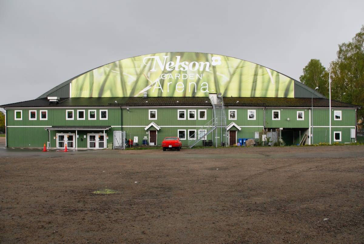 En stor grön byggnad med en banner som annonserar "Nelson Garden Arena" syns under en gråmulen himmel. Framför byggnaden finns en grusplan och en röd bil är parkerad. Till vänster i bild syns två koner och fler träd till höger.