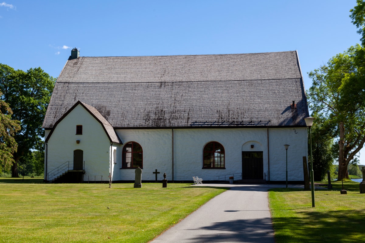 En vit kyrkobyggnad med grått tak ses rakt framifrån under dagtid, omgiven av gräsmatta och träd. En asfalterad gång leder mot kyrkans ingångar. Kyrkan har flera fönster och dörrar samt en mindre utskjutande del på vänster sida. Två gravstenar syns.