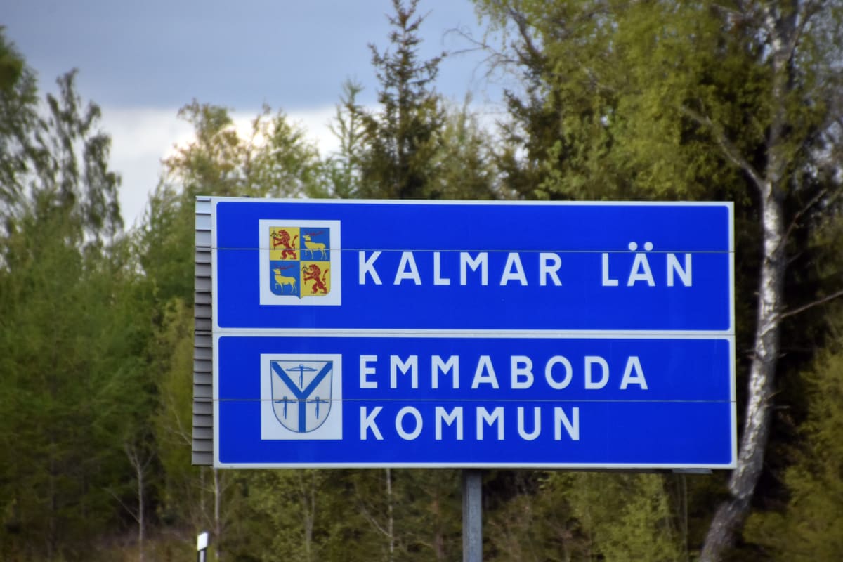 En blå skylt med vita bokstäver visar "KALMAR LÄN" och "EMMABODA KOMMUN". Skylten är monterad på en stolpe och omges av gröna träd under en delvis molnig himmel.