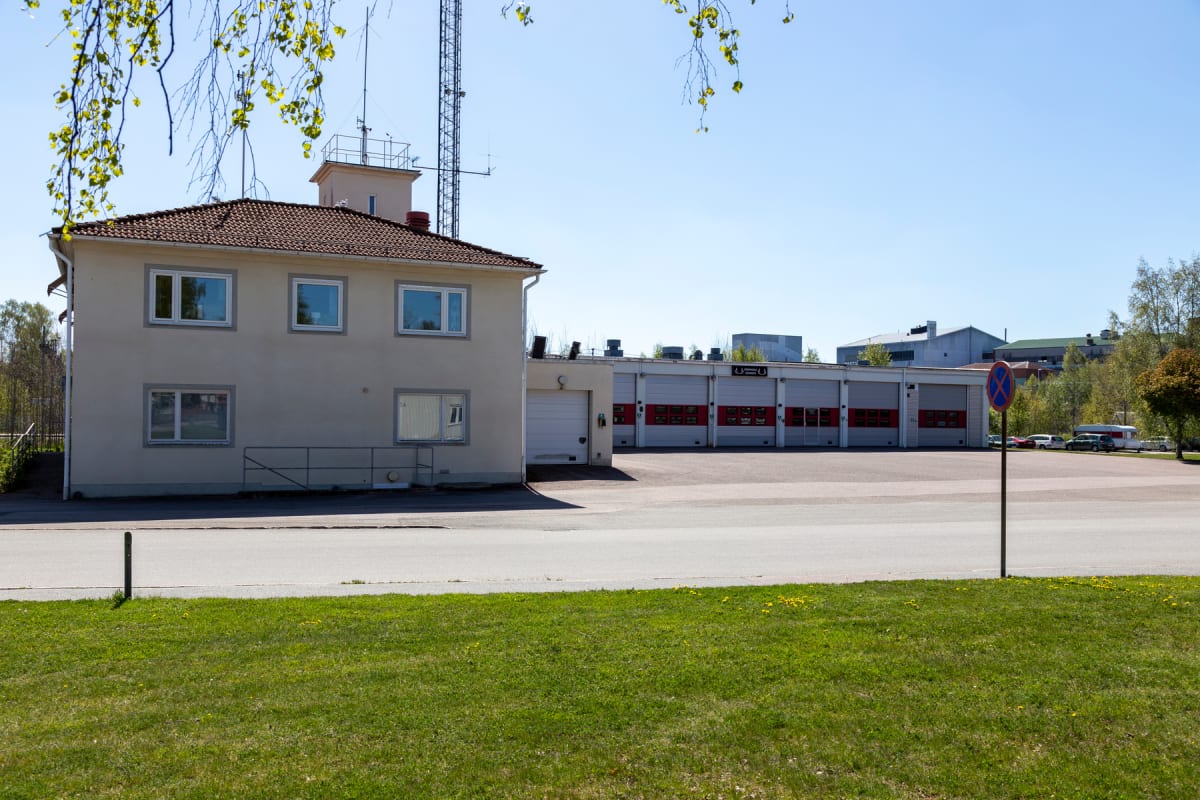En beige byggnad med tegeltak och flera fönster står till vänster, med en rad med garageportar med röda markeringar till höger. I bakgrunden syns skog, andra byggnader och fordon. Framför grön gräsmatta och asfalt, med en skylt med ett rött kryss öve