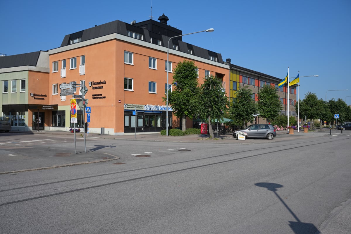 En gathörna med byggnader på vänster sida och vägkorsning framför. En orange byggnad med skylten "Emmaboda kommun" och "Besökscenter & Turistinformation" dominerar. Träd, parkerade bilar och flaggstänger med svenska flaggor syns.