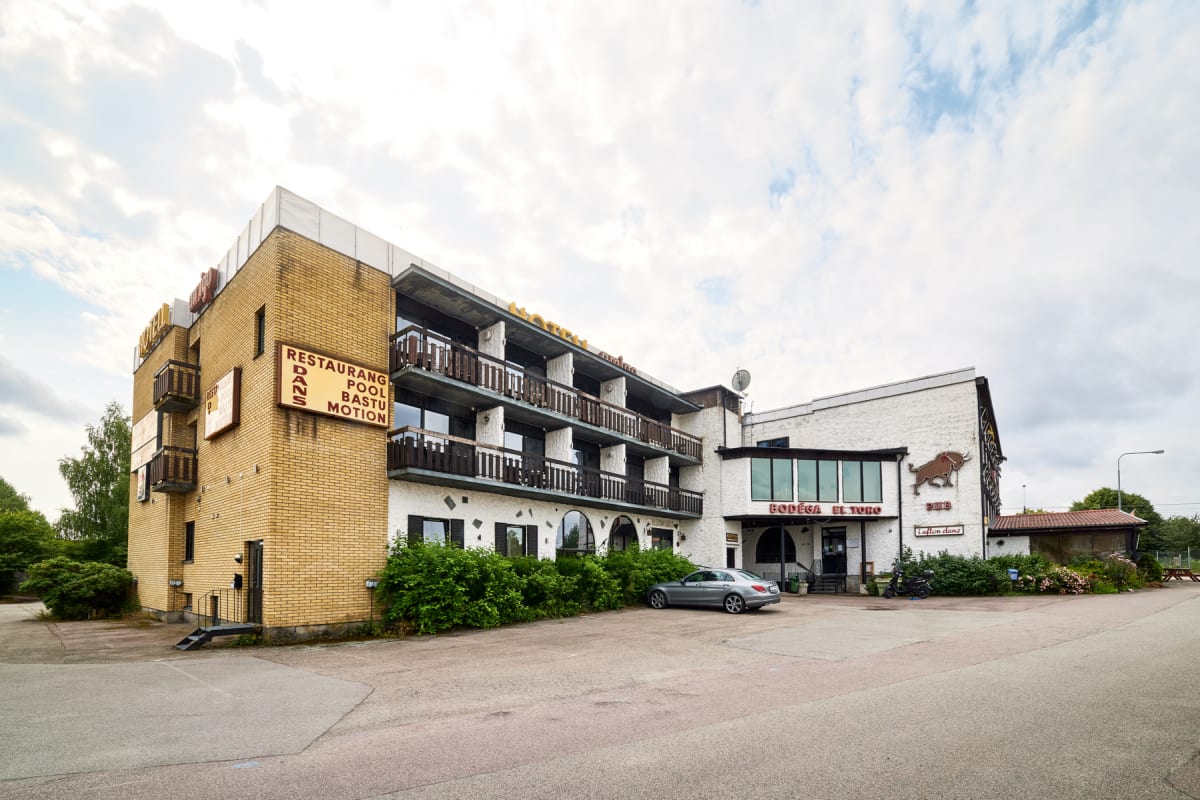 En stor byggnad med gul tegelfasad och vit, putsad del under en molnig himmel. Byggnaden har skyltar med text som 'RESTAURANG', 'POOL', 'BASTU', 'DANS' och 'MOTION', samt 'BODEGA EL TORO PUB'. En grå bil står parkerad framför entrén.