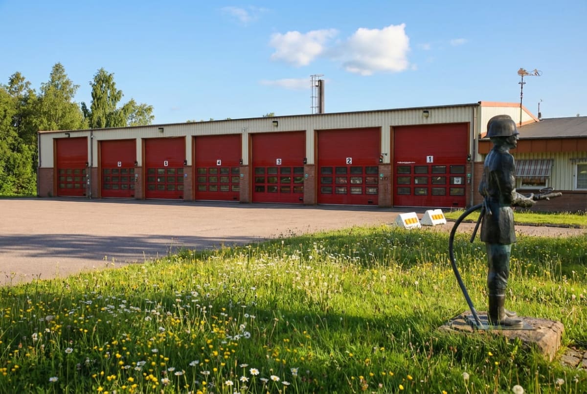 En bild som visar en brandstation med sex röda portar, numrerade från 1 till 6, under en klarblå himmel med några moln. Framför stationen finns en asfalterad yta och en gräsmatta med vita och gula blommor. En staty av en brandman syns i förgrunden.