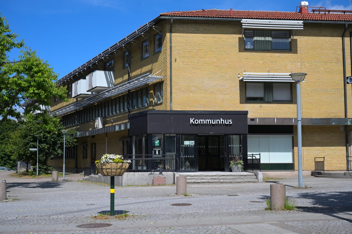 En gul tegelbyggnad med texten "Kommunhus" över entrén, belägen vid en kullerstensbelagd yta. Gröna träd och växter syns runt byggnaden under en klarblå himmel. Ett blomsterarrangemang står på en stolpe framför entrén.