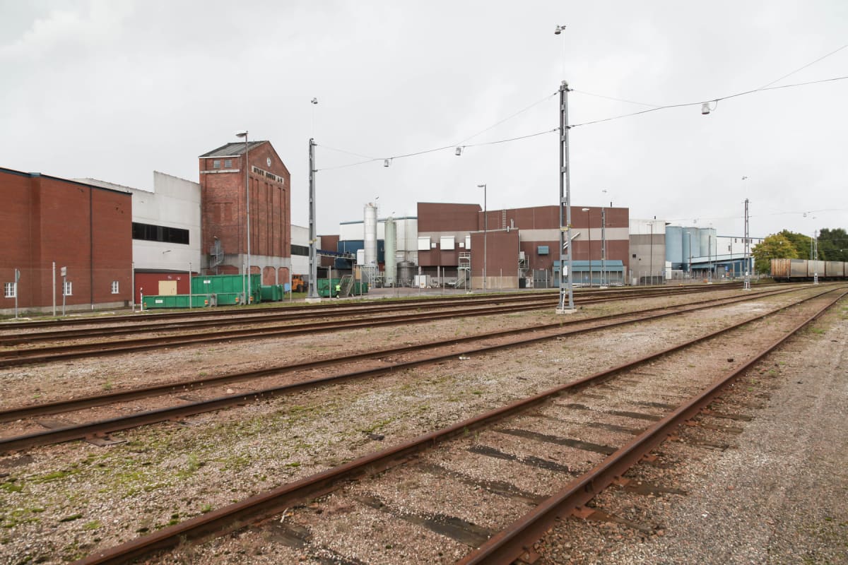 Bilden visar ett industriområde med flera byggnader och järnvägsspår i förgrunden. Till vänster syns en röd tegelbyggnad med texten "NYBYGGNADSMEDEL" och nedanför, flera gröna containrar. I mitten och till höger finns grå och bruna lagerbyggnader med