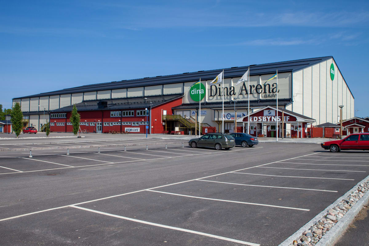En stor byggnad, delvis röd och delvis vit med mörk tak, syns under en molnfri blå himmel. Utanför byggnaden finns en asfalterad parkeringsplats med vita markeringar. Flera bilar är parkerade. Flaggstänger med flaggor står framför entrén.