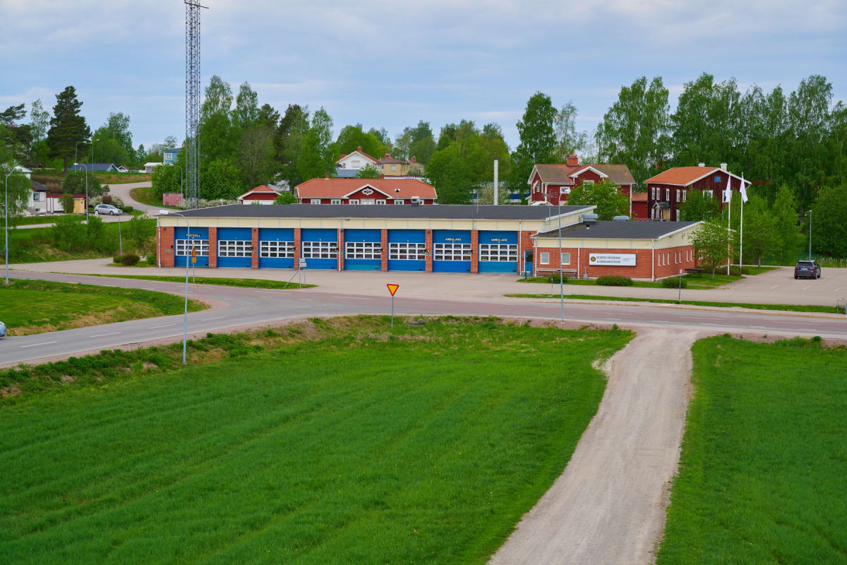 En översiktsbild av en brandstation med flera blå garageportar, omgiven av grönskande träd och gräsmattor. En asfalterad väg löper i förgrunden, och i bakgrunden syns bostadshus med röda tak under en molnig himmel.