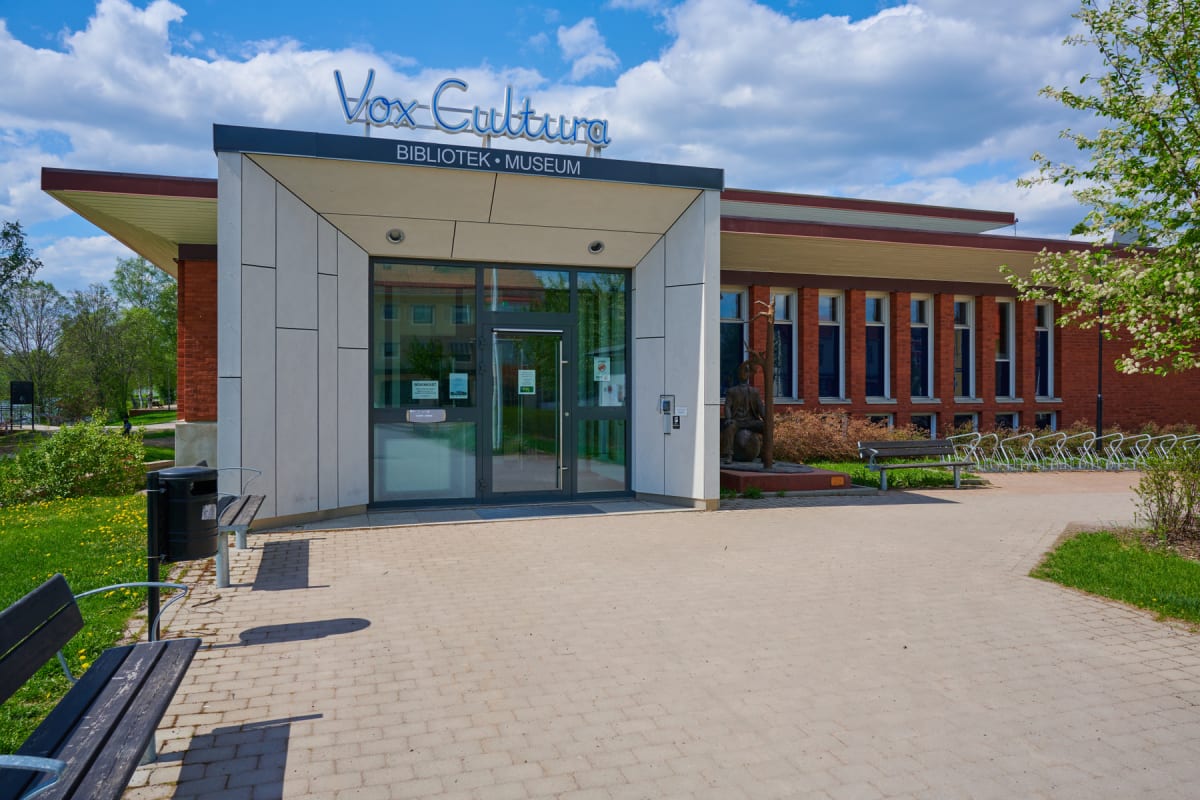 En fasad på en byggnad med texten "Vox Cultura BIBLIOTEK • MUSEUM" syns under en blå himmel med vita moln. Framför ingången finns en belagd yta, gräsmattor, bänkar, en papperskorg, cykelställ och en staty.
