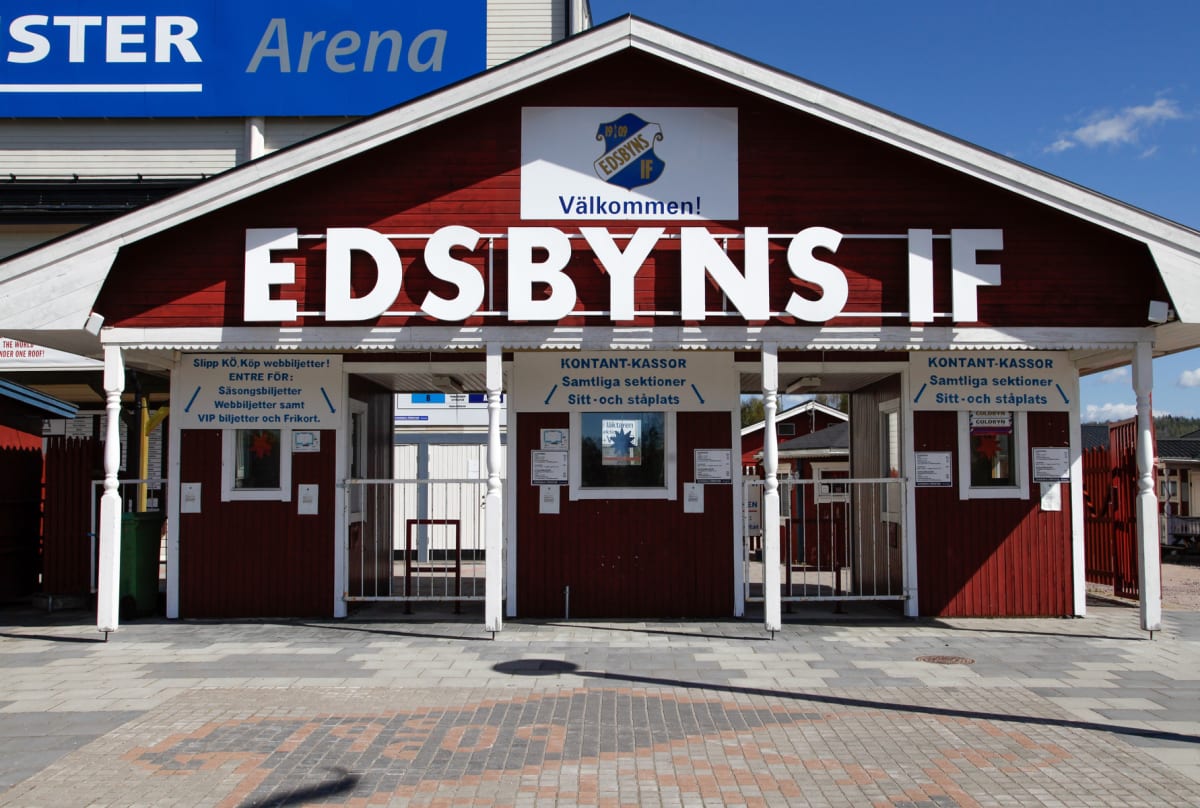 En röd byggnad med vita detaljer och skylten "EDSBYNS IF" är synlig framför en större vit byggnad märkt "STER Arena". Byggnaden har tre öppningar med kassor och grindar. På marken framför byggnaden finns ett mönstrat stenläggningsarbete.
