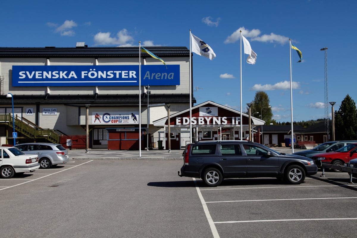 En utomhusbild som visar byggnader med skyltar för "Svenska Fönster Arena" och "Edsbyns IF" under en delvis molnig blå himmel. Framför byggnaderna finns en parkeringsplats med flera parkerade bilar och flaggstänger med flaggor.