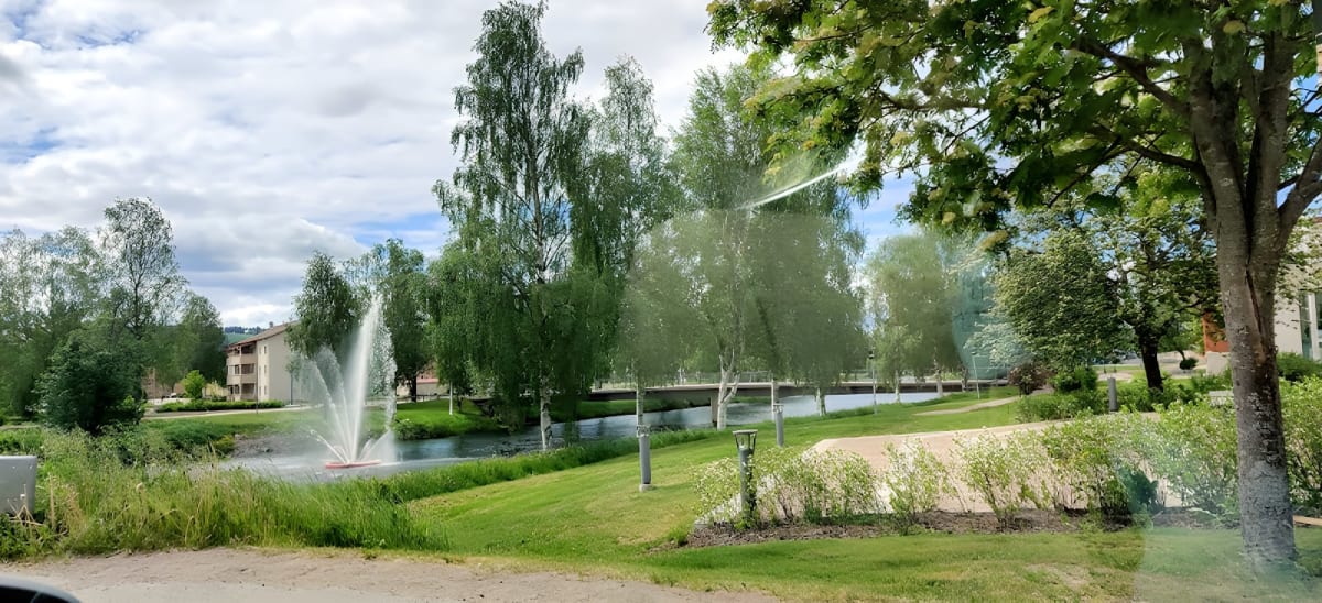 En utomhusbild som visar en fontän i en vattenyta omgiven av gräs och lummiga träd, under en molnig himmel. I bakgrunden syns bostadshus och en bro.