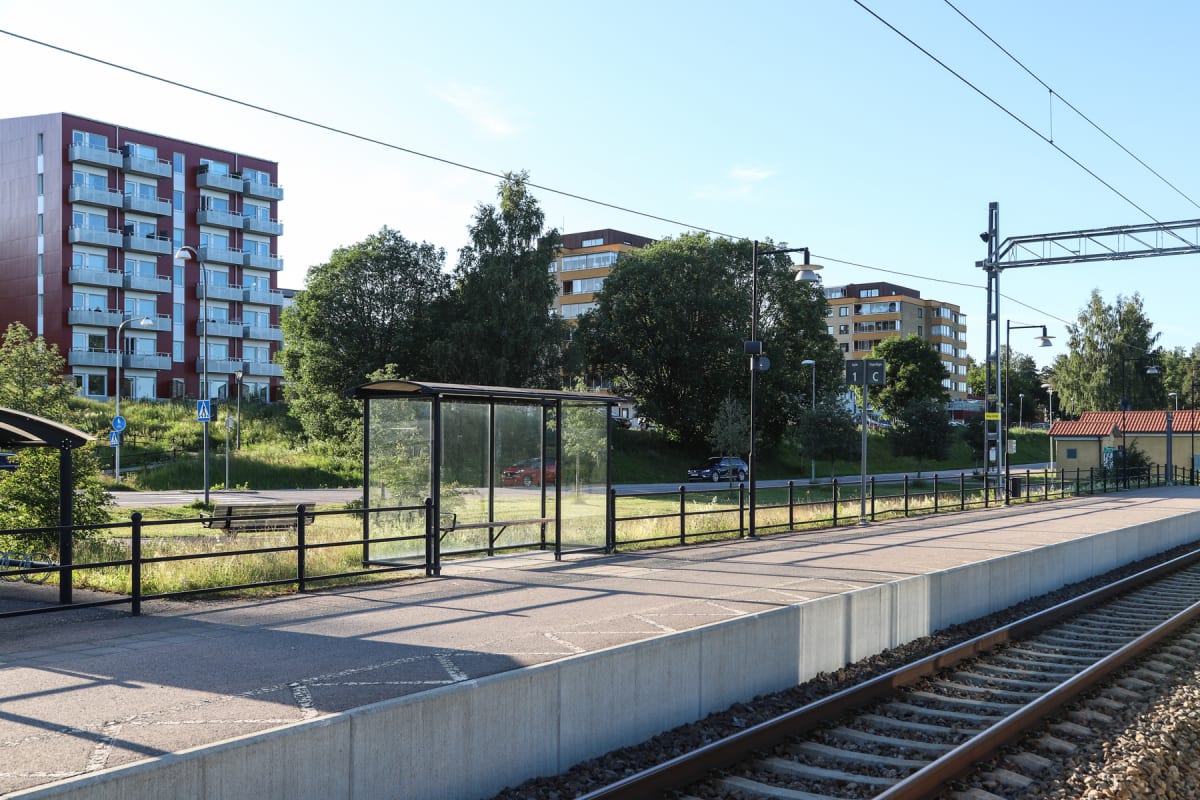 En bild av en tågperrong med en busskur och järnvägsspår i förgrunden. I bakgrunden syns flerbostadshus omgivna av träd och grönska, samt en väg med ett par bilar. Himmelen är klarblå.