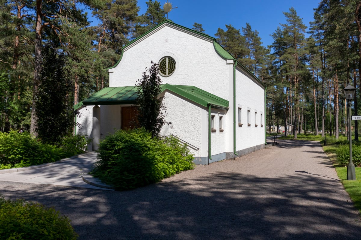 En vit byggnad med grönt tak och gröna detaljer, omgiven av tallar och grönskande buskar under en klarblå himmel. Framför byggnaden finns en grusväg och en stenlagd gång.