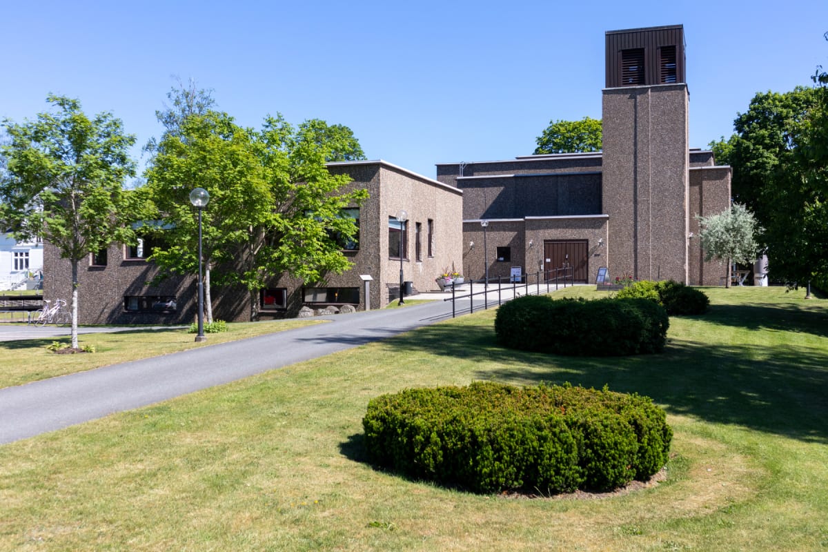 En modern kyrkobyggnad av betong med ett kvadratiskt klocktorn syns under en klarblå himmel. Framför kyrkan finns en asfalterad gångväg, gräsmattor och lövträd, samt buskar på tomten. På gångvägen står även en lyktstolpe.