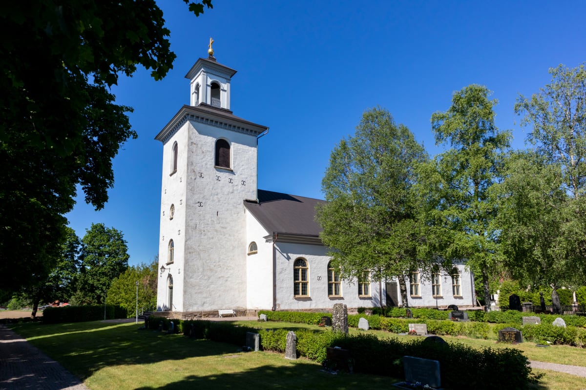 En vit kyrka med ett torn med ett gyllene kors på toppen, syns under en klarblå himmel. Runt kyrkan finns gräsmattor, träd och gravstenar. Solen lyser och skapar skuggor på marken.