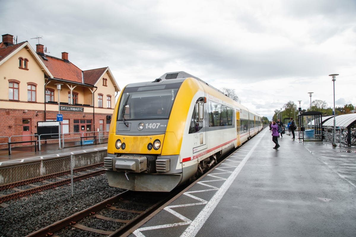 En gul och vit motorvagn står vid en perrong framför en tågstation med ett tegelrött tak och en skylt där det står 'Skillingaryd'. Himmel är molnig och perrongen är våt, vilket kan indikera att det nyligen regnat.