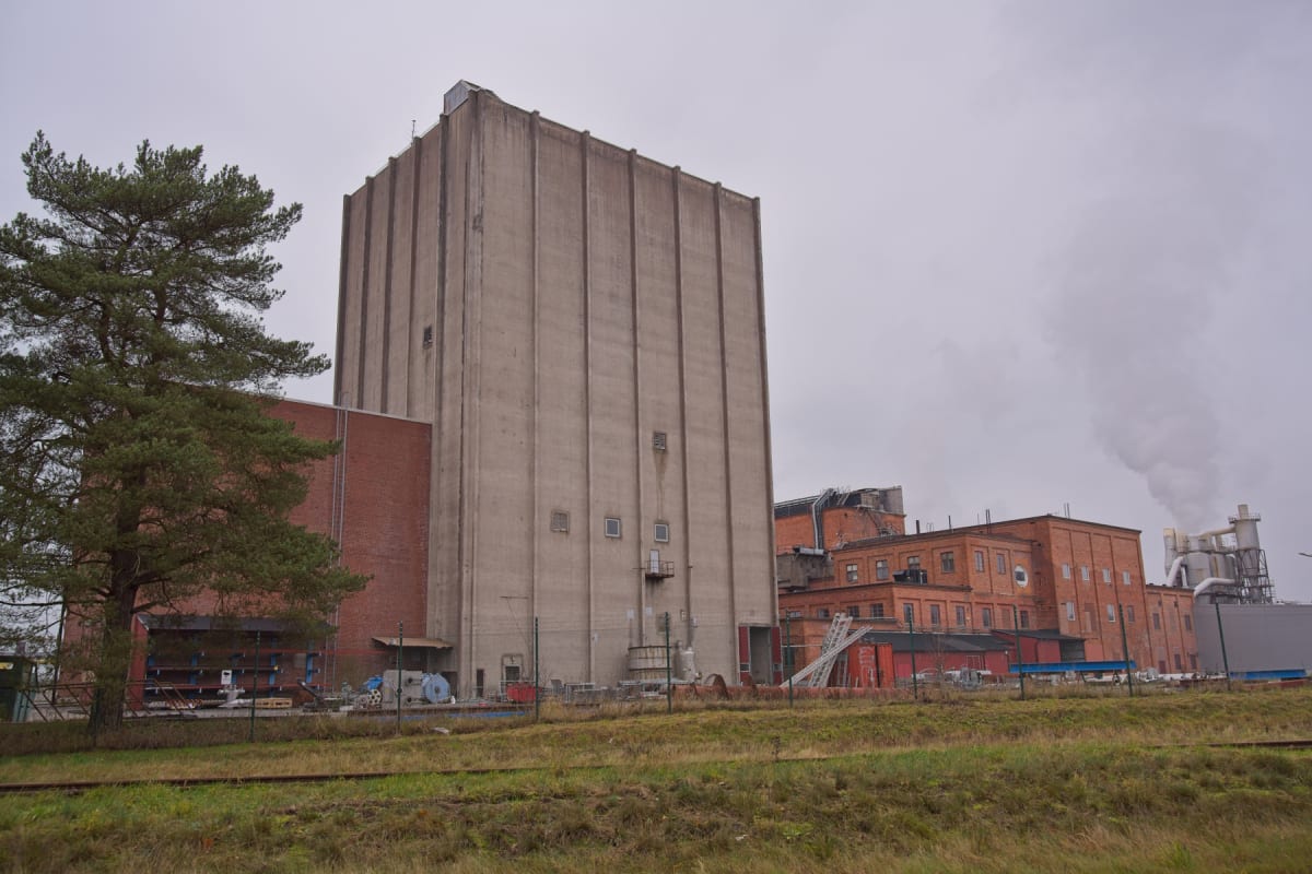 En bild visar en industriell anläggning under en molnig himmel. I förgrunden syns gräs och vad som ser ut som tågräls. Centralt i bilden dominerar en stor, grå betongbyggnad, flankerad av rödare tegelbyggnader. Till vänster står ett barrträd, och i b