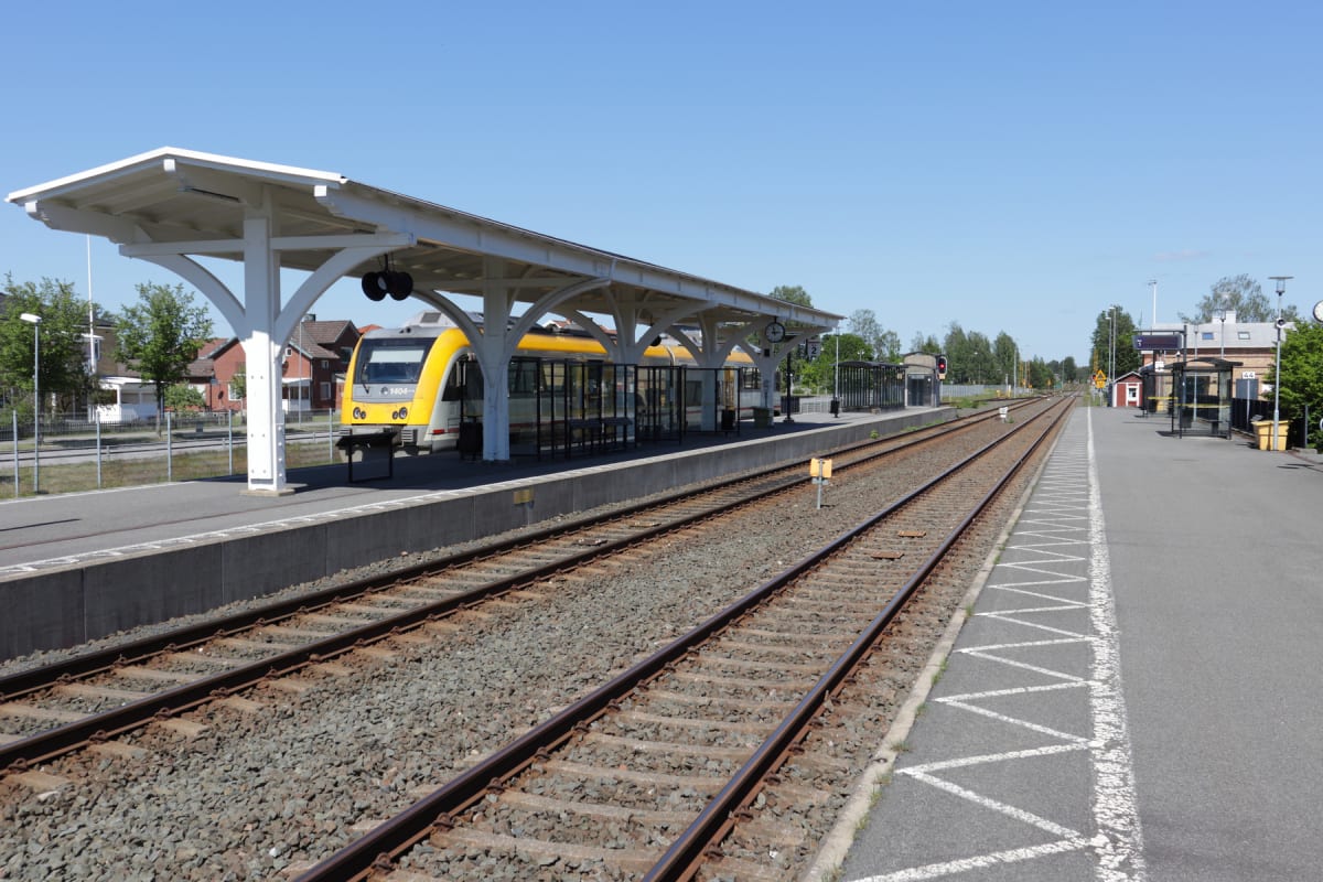 En gul och vit pendeltåg står vid en plattform på en tågstation under en solig dag. Två järnvägsspår löper parallellt med plattformen, och en skyddad väntplats med ett vitt tak syns till vänster.