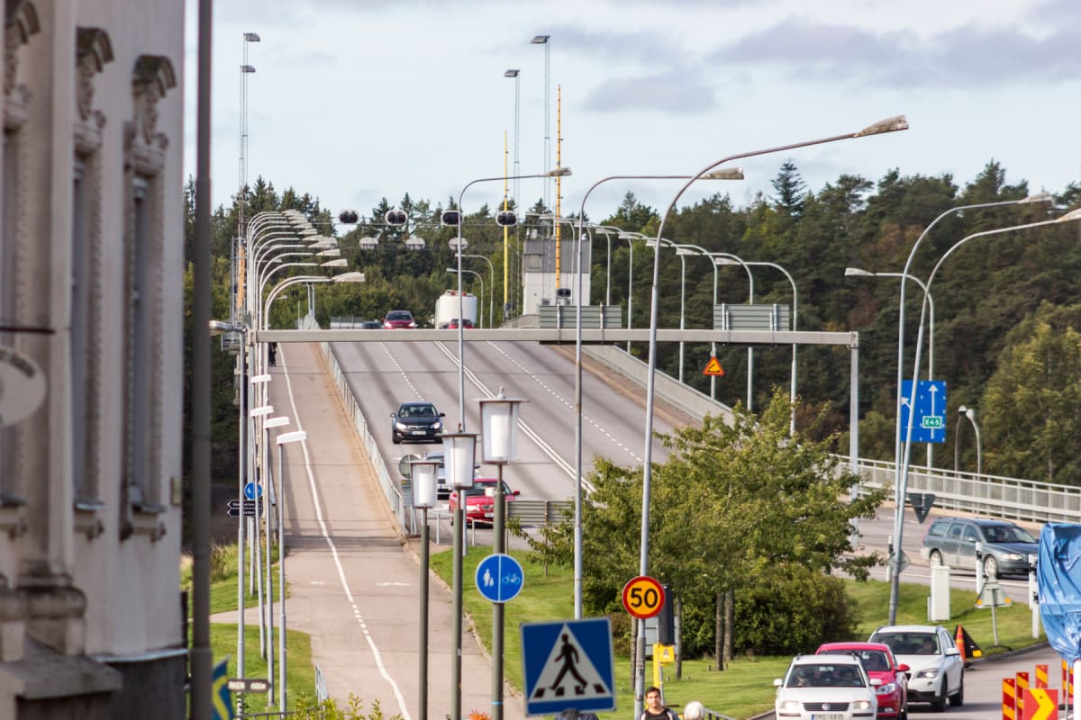 En bro i dagsljus som sträcker sig över marken med tallskog i bakgrunden. En körbana med flera bilar är central i bilden. Längs vänsterkanten syns delar av en byggnad. Längs bron finns flera lampstolpar och trafikskyltar. Två personer skymtar i nedre