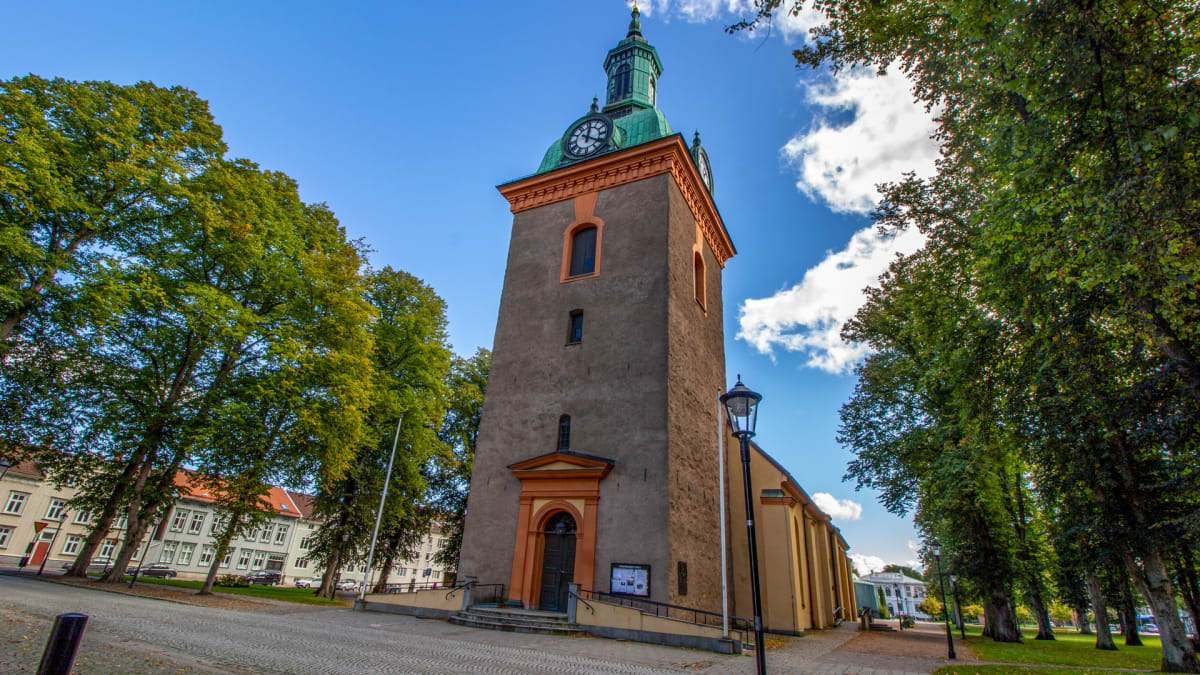 En vertikal bild av en grå kyrkobyggnad med grönbrun kopparkupol och klocka, omgiven av höga, lummiga träd. Nedanför kyrkan syns en kullerstensbelagd yta och till vänster om den avtecknar sig delar av byggnader.