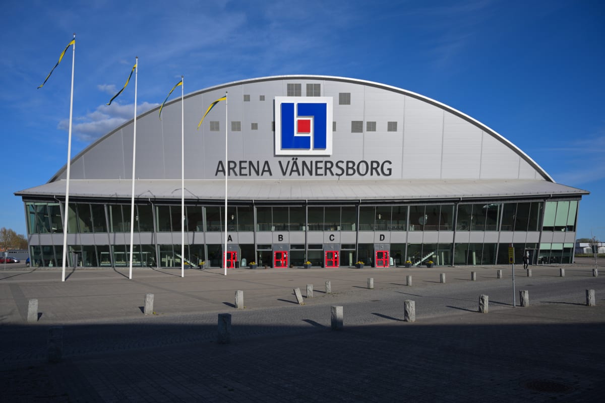 En modern arena med grå fasad och stora glaspartier under en blå himmel. Framför arenan finns en stenlagd yta med flera flaggstänger med flaggor i gult och blått, samt stenpollare. Byggnaden har texten "ARENA VÄNERSBORG" och en logotyp.