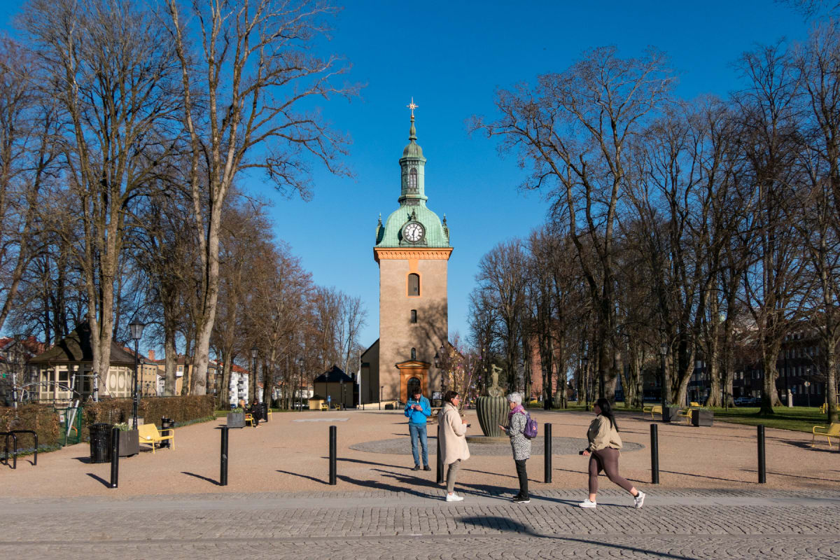 En stor översiktsbild av ett torg eller en öppen plats med en kyrktorn i mitten och kala träd på båda sidor. På torget syns flera personer, några bänkar och lyktstolpar. Himmelen är klarblå.
