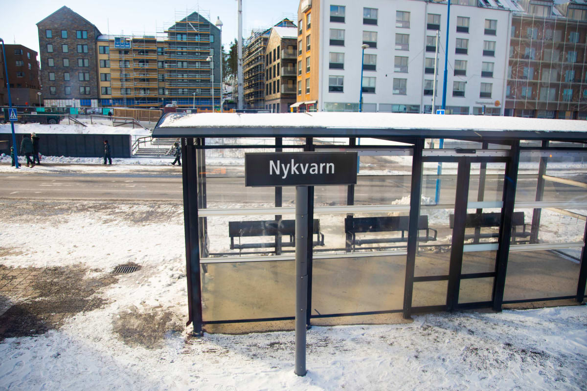 En gata täckt av snö i förgrunden, med en busskur med skylten "Nykvarn" i centrum. I bakgrunden syns flera byggnader, varav några är under konstruktion, samt människor som går längs gatan.
