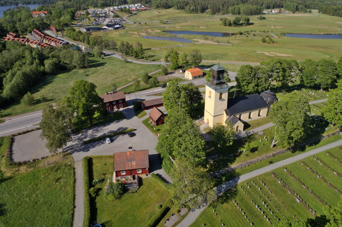 Flygfoto över ett landskap med en gul kyrka med klocktorn och en omgivande kyrkogård med gravstenar. I förgrunden syns flera röda byggnader. En väg löper genom landskapet, och i bakgrunden finns en golfbana med två vattenhinder samt en sjö och skog.