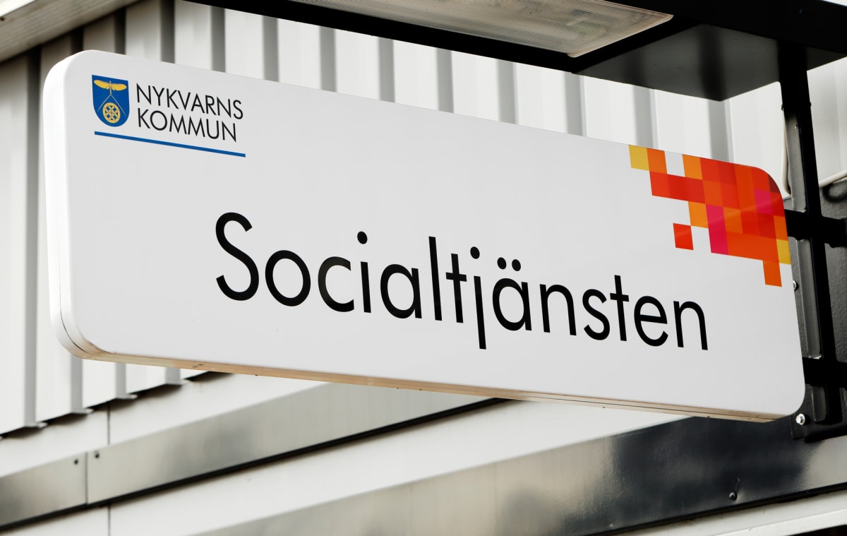 En vit skylt med texten "Socialtjänsten" i svarta bokstäver och logotypen för Nykvarns kommun i övre vänstra hörnet, hänger under ett tak och framför en vit vägg.