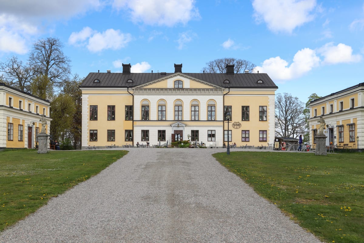 En stor, gul byggnad med vita detaljer och svarta tak dominerar mitten av bilden, flankerad av två mindre liknande byggnader på sidorna. En grusgång leder fram till huvudbyggnadens entré, omgiven av gräsmattor med viss vårblomning. Flera personer sit