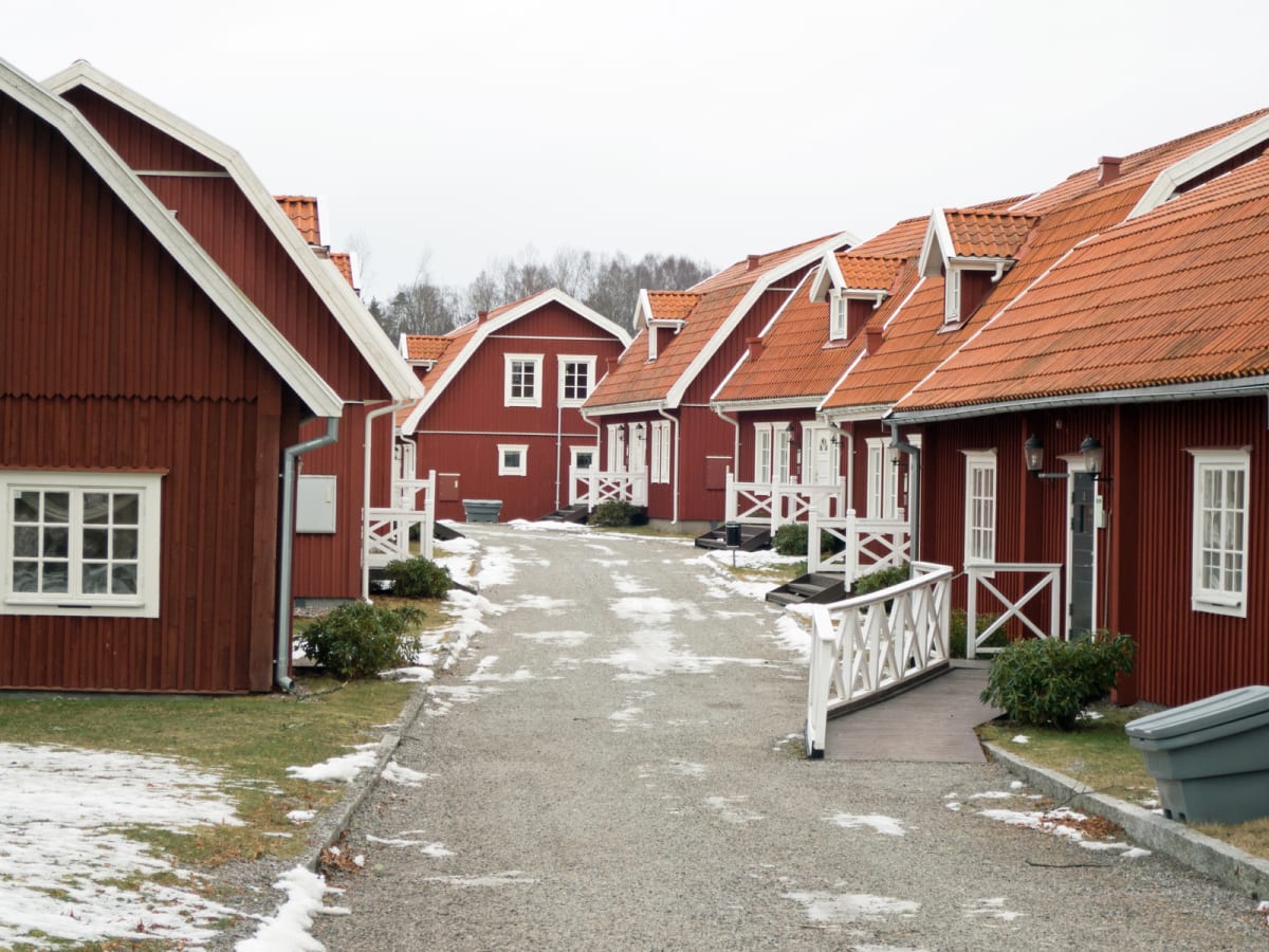 En gata kantad av röda hus med vita detaljer och orange tak. Marken är grusad med fläckar av snö, och gräsmattorna är delvis snötäckta. Himlen är grå och träden i bakgrunden saknar löv. Ett grått kärl syns i bildens nedre högra hörn.