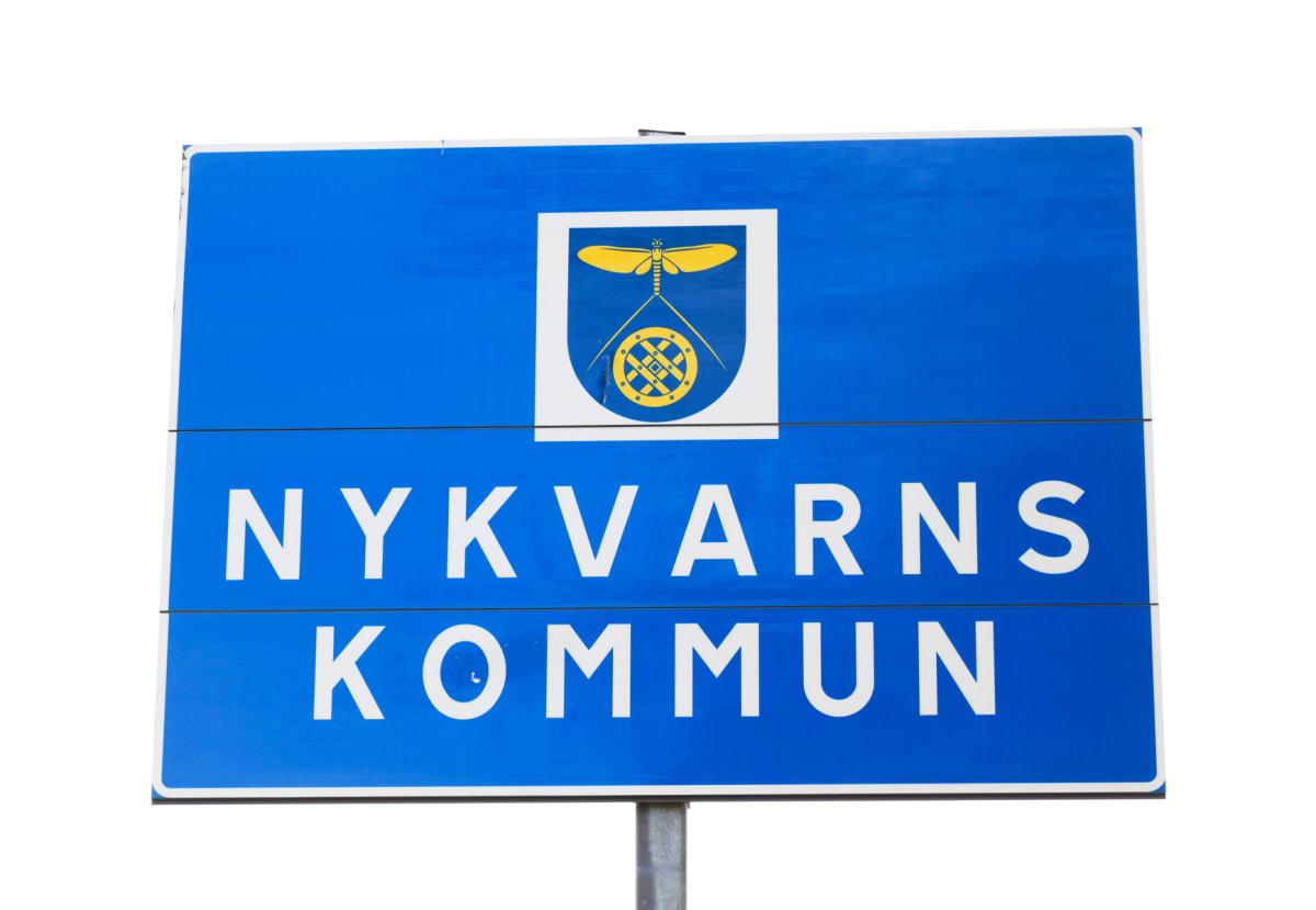 En blå skylt med texten "NYKVARNS KOMMUN" i vit färg, samt ett kommunvapen i gult och vitt. Skylten är monterad på en stolpe och har vita kanter. Bakgrunden är vit.