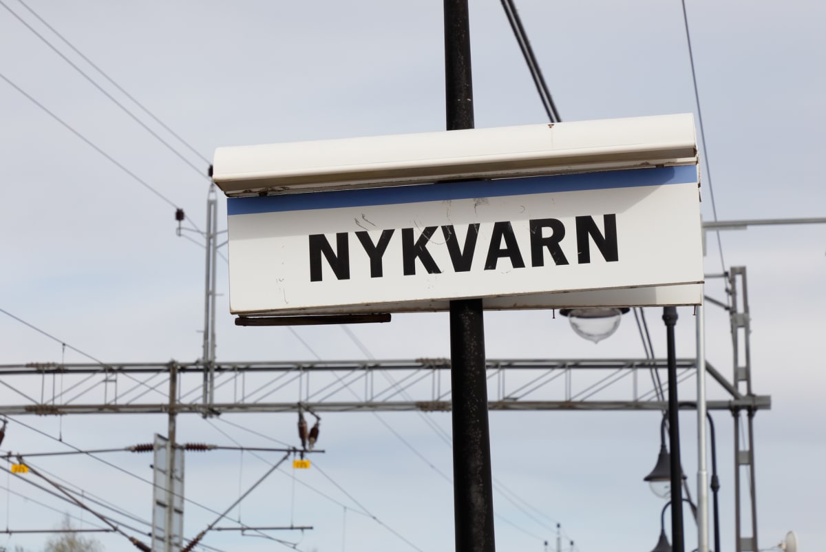 En vit skylt med en blå rand och texten "NYKVARN" i svarta versaler. Skylten är monterad på en svart stolpe. I bakgrunden syns kraftledningar, elstolpar och delar av en lyktstolpe under en ljus himmel.