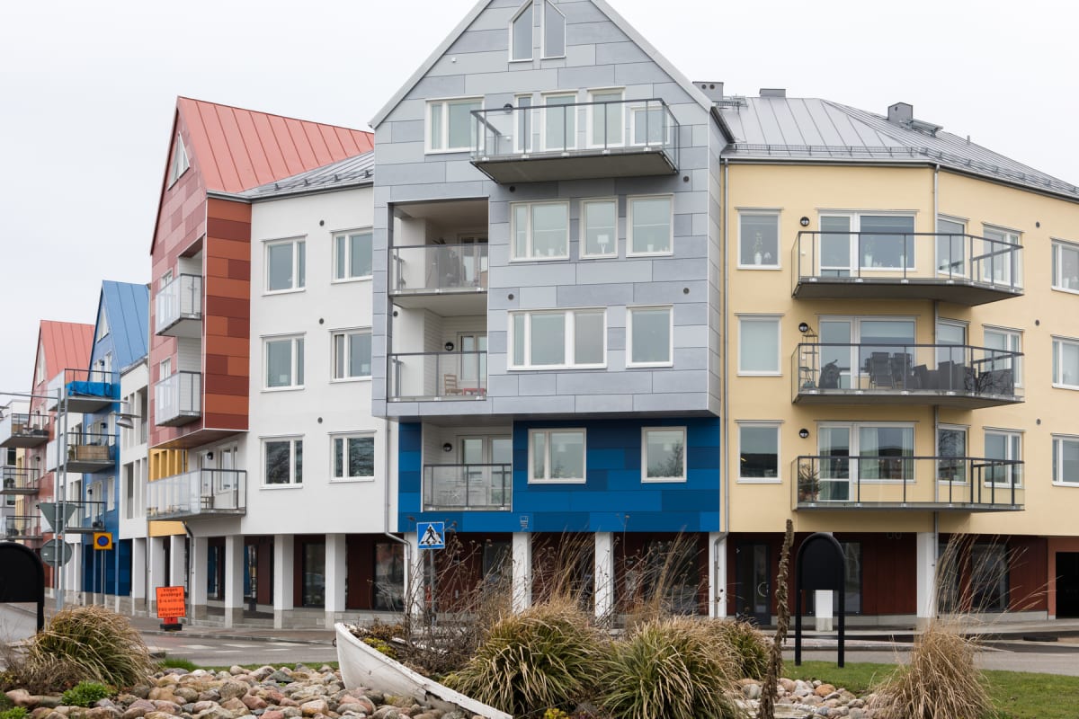 En bild utomhus som visar en rad flerbostadshus i olika färger, med flera balkonger. I förgrunden syns en liten odlingsyta med stenar, gräs och en ommålad gammal båt. En skylt med “Vägen ombyggd, följ orange vägvisning” är synlig.