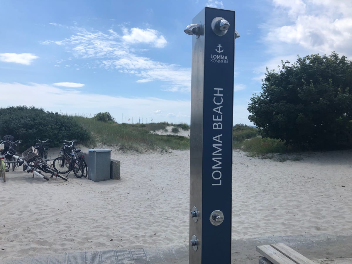 En vertikal, mörkblå pelare med texten "LOMMA KOMMUN" och "LOMMA BEACH" står på en sandstrand under en ljusblå himmel med moln. I bakgrunden syns sanddyner med gräs och gröna buskar, samt flera cyklar och en papperskorg.
